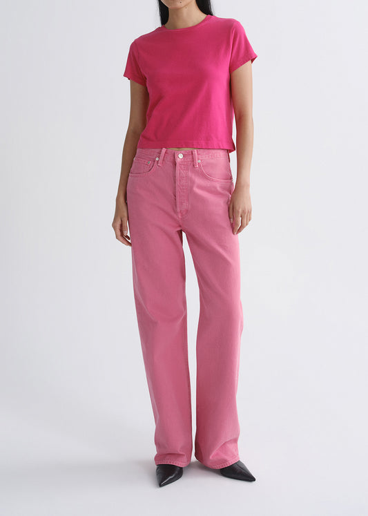 A model wearing a bright pink short-sleeve crewneck t-shirt and matching pink wide-leg jeans, paired with black pointed-toe heels, against a grey background.
