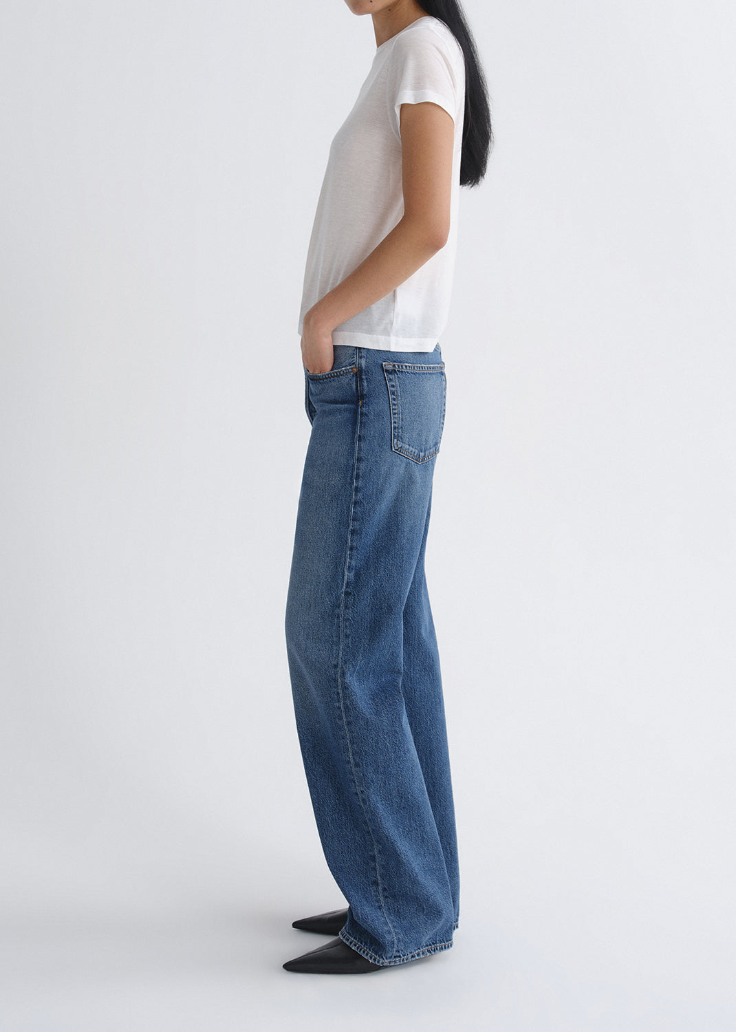 Person wearing a white short sleeve crewneck tee with medium wash wide leg jeans viewed from the side on a grey background.