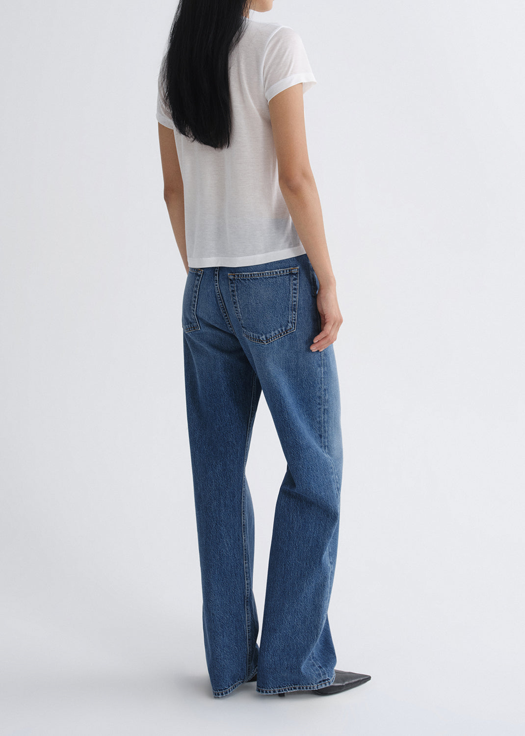 Person wearing a white short sleeve crewneck tee with medium wash wide leg jeans viewed from the back on a grey background.