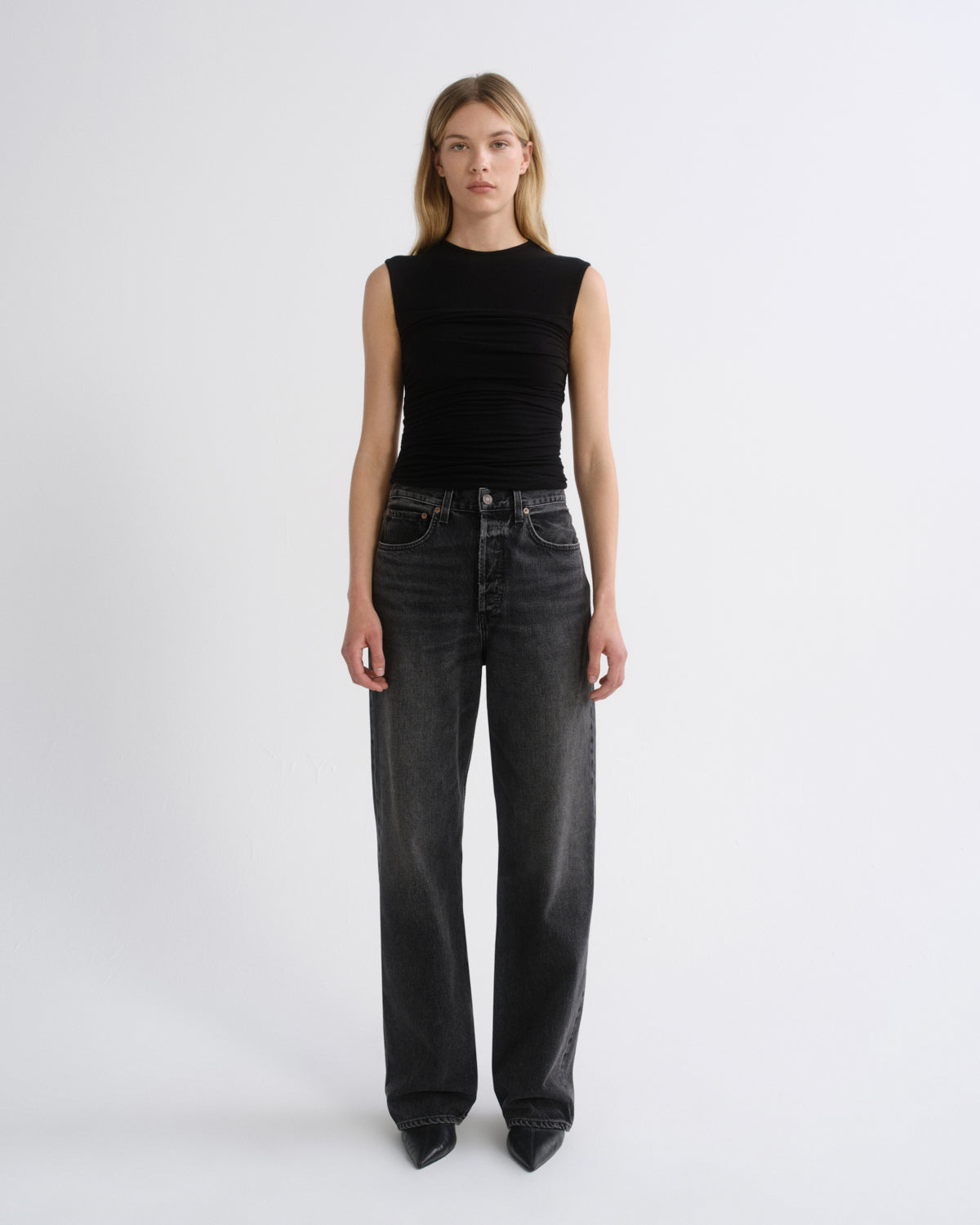 A model stands against a plain light gray studio background wearing a sleeveless black ribbed top and dark gray wide-leg jeans.