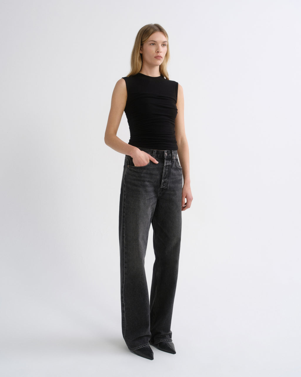 A model stands against a light gray studio background wearing a sleeveless black ribbed top and dark gray wide-leg jeans.