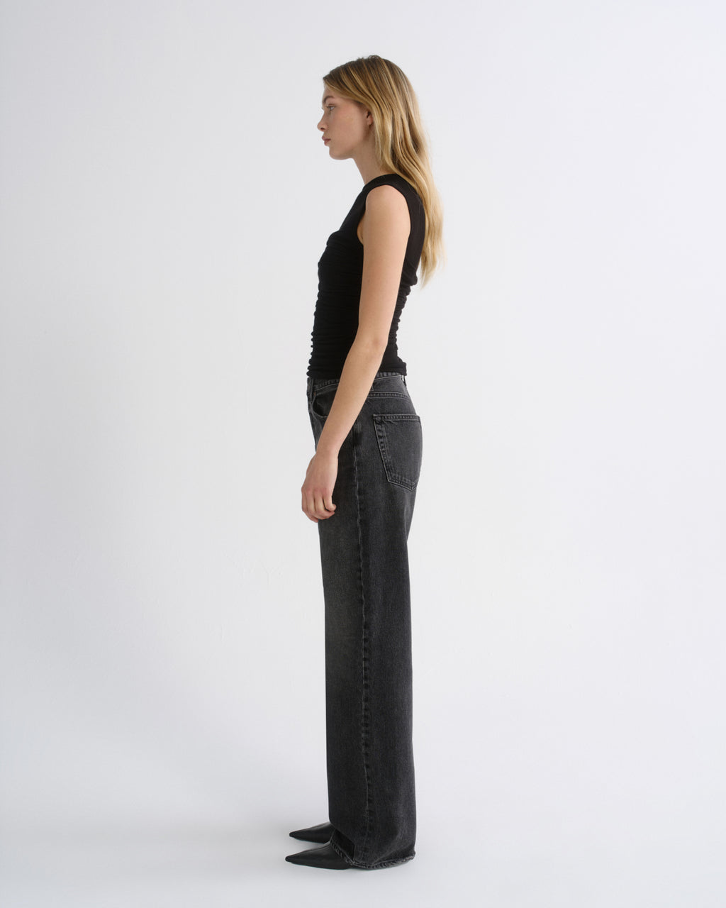 A woman stands in profile wearing a black sleeveless top and dark gray wide-leg jeans against a plain light gray studio background.