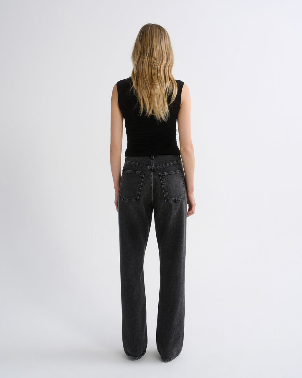 A woman viewed from behind wearing a black sleeveless top and dark charcoal gray wide-leg jeans against a plain light gray studio background.