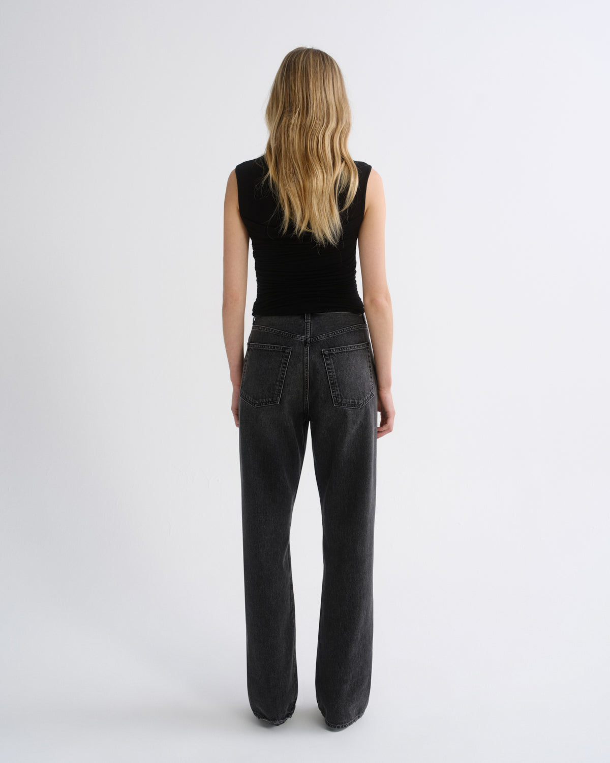 A woman viewed from behind wearing a black sleeveless top and dark charcoal gray wide-leg jeans against a plain light gray studio background.