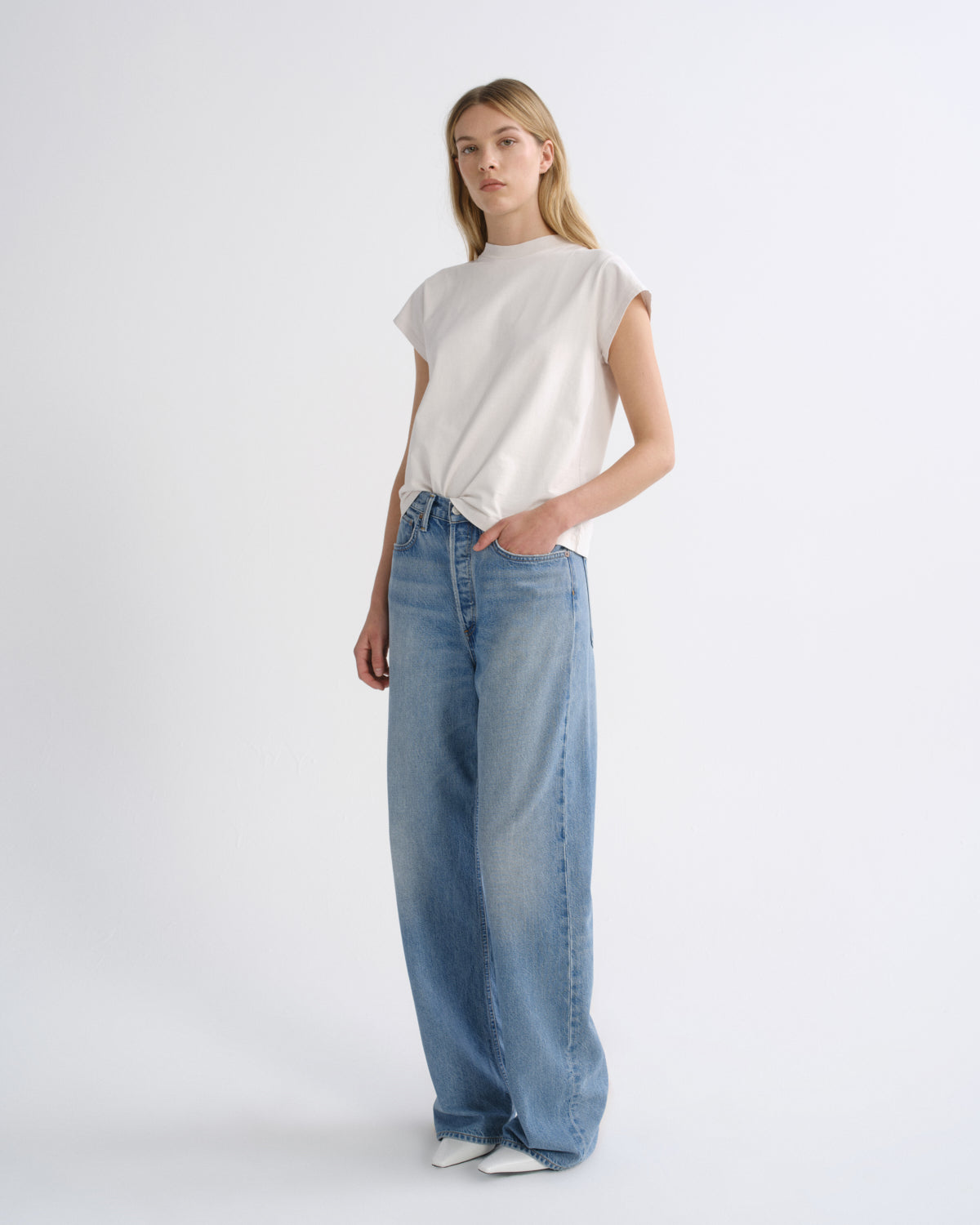 A woman modeling a white short-sleeve t-shirt tied at the waist with wide-leg light blue denim jeans, standing against a plain white studio background.