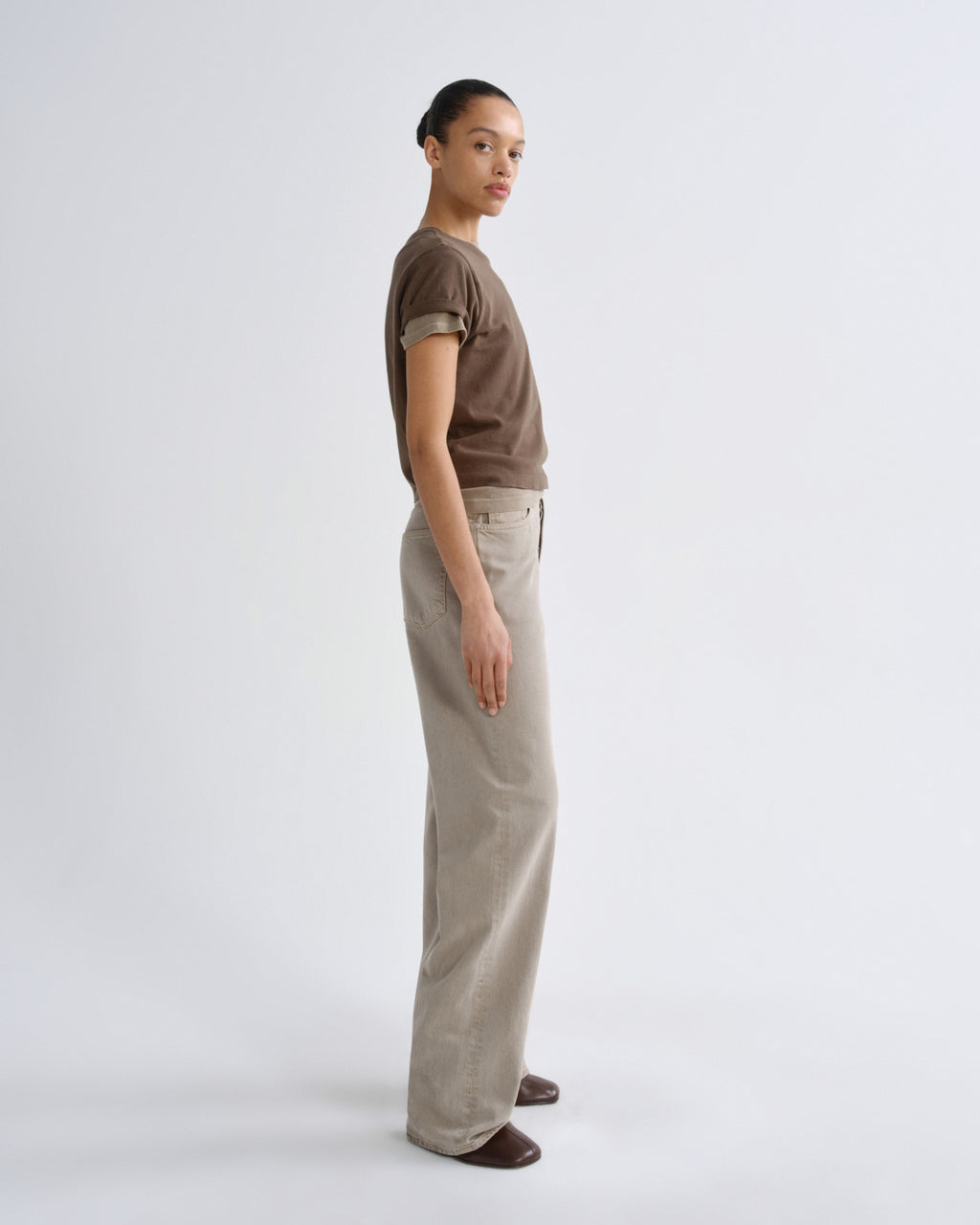 A model stands in profile wearing a brown short-sleeve t-shirt and light beige wide-leg trousers against a plain light gray studio background.