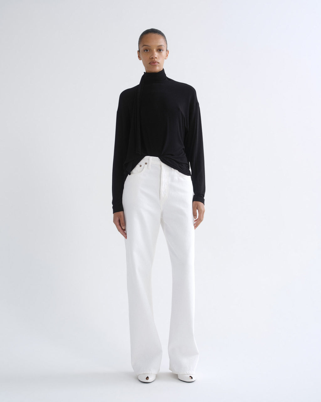 A model stands against a plain white studio background wearing a black long-sleeve turtleneck top and high-waisted white wide-leg trousers.