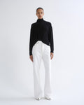 A model stands against a plain white studio background wearing a black long-sleeve turtleneck top and high-waisted white wide-leg trousers.