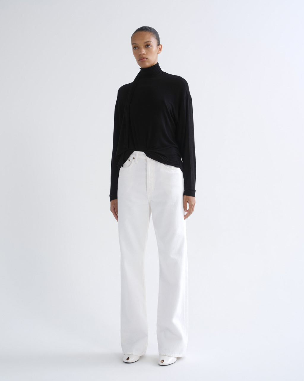 A model stands against a plain white studio background wearing a black long-sleeve turtleneck top and high-waisted white wide-leg trousers.
