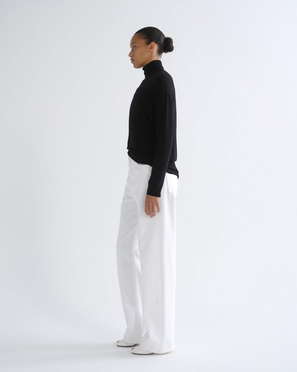 A model shown in profile wearing a black long-sleeve mock neck top and white wide-leg trousers against a plain white studio background.