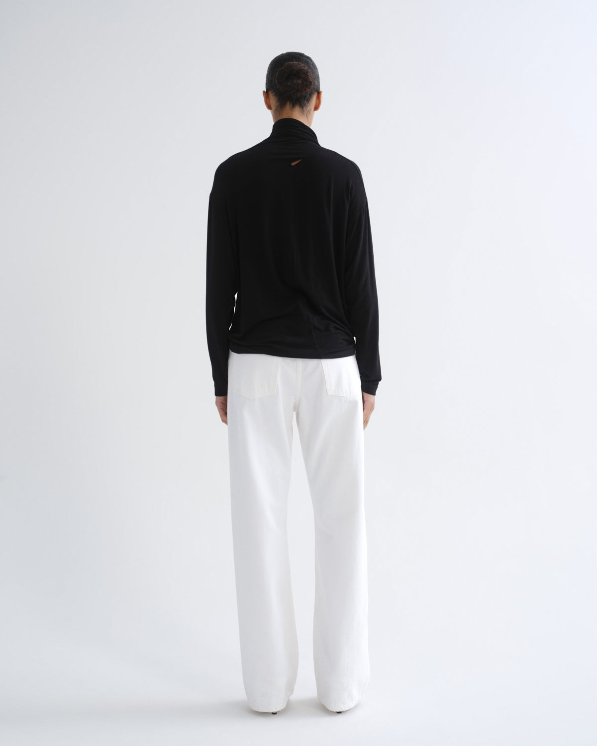 A model viewed from the back wearing a black long-sleeve mock neck top and white wide-leg trousers, standing against a plain light gray studio background.