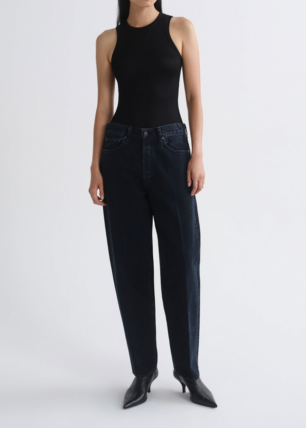 A model wearing a black sleeveless bodysuit, dark blue straight-leg jeans, and black pointed-toe heeled boots, standing against a grey background.