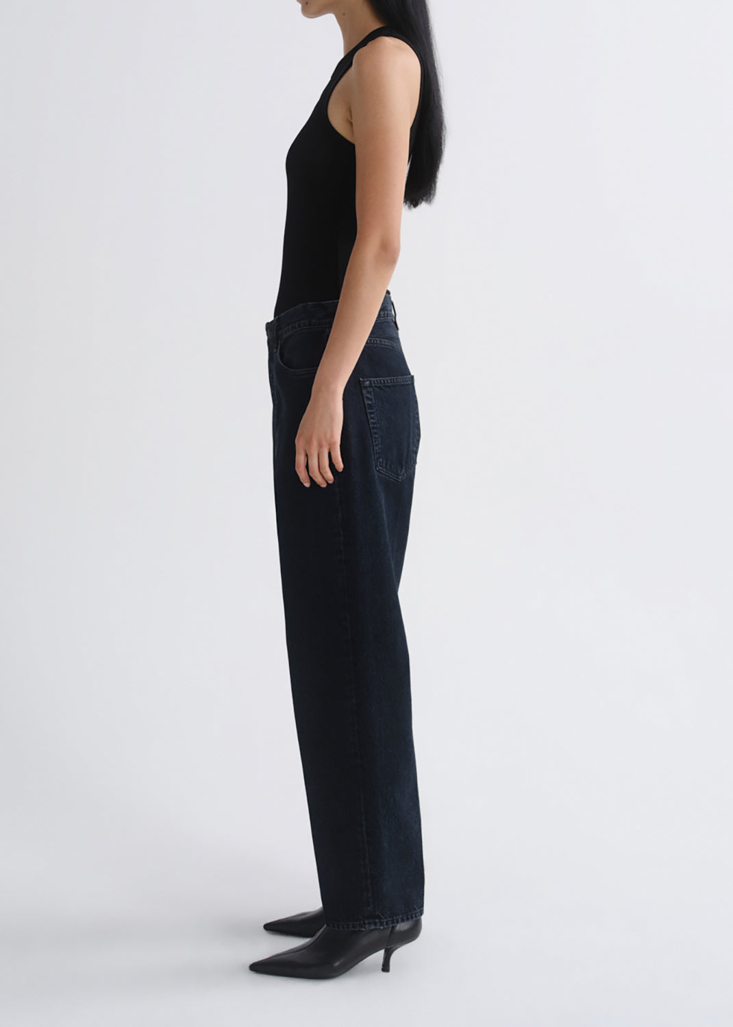 A model shown from the side wearing dark wash wide-leg jeans, a black sleeveless top, and black pointed-toe heeled boots against a grey background.