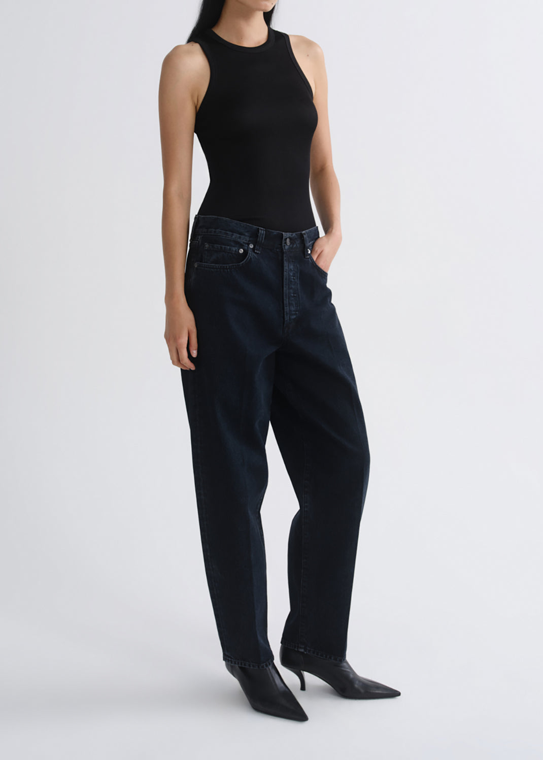A model wearing dark wash wide-leg jeans, a black sleeveless top, and black pointed-toe heeled boots, standing against a grey background.