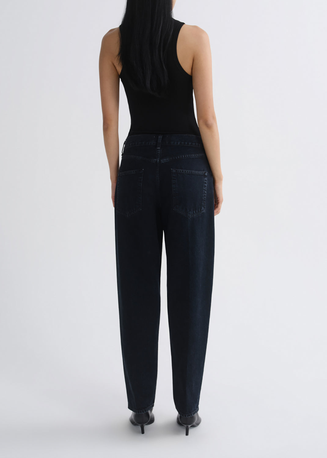 A woman shown from the back wearing dark wash wide-leg jeans, a black sleeveless top, and black heels against a grey background.