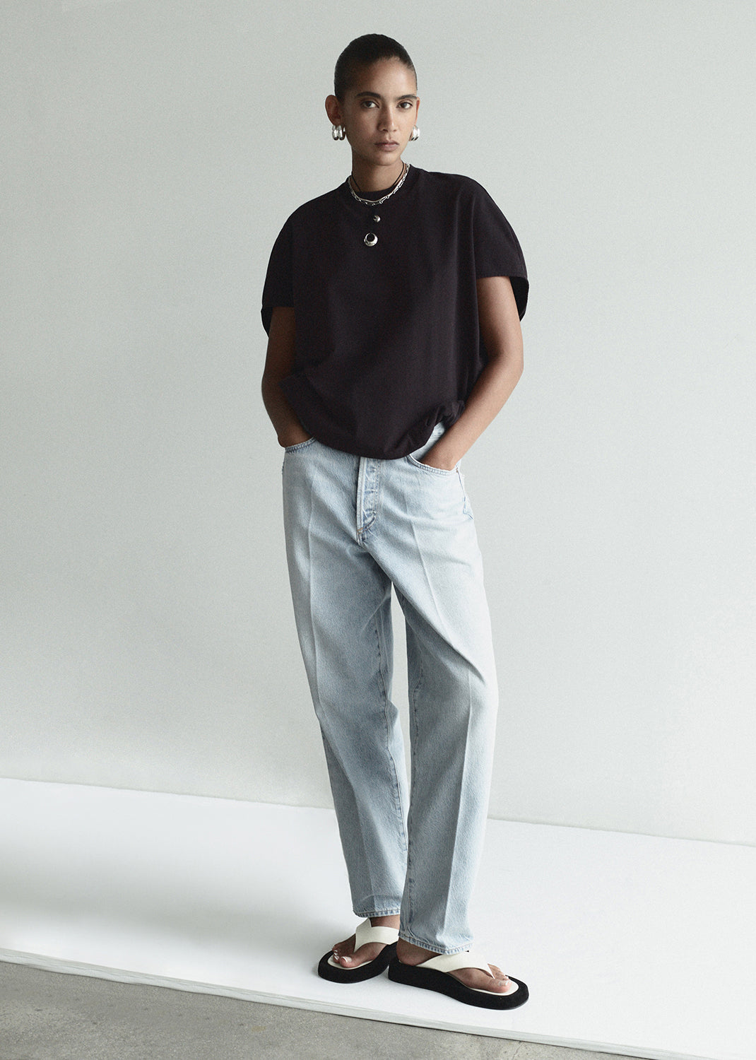 Person wearing a black t-shirt and light blue jeans standing against a white wall.