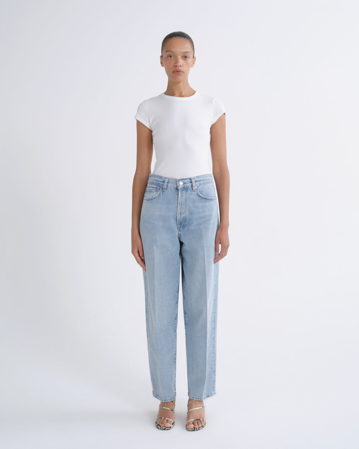 A model stands against a light gray studio background wearing a white short-sleeve top and light-wash blue straight-leg jeans with beige strappy heels.