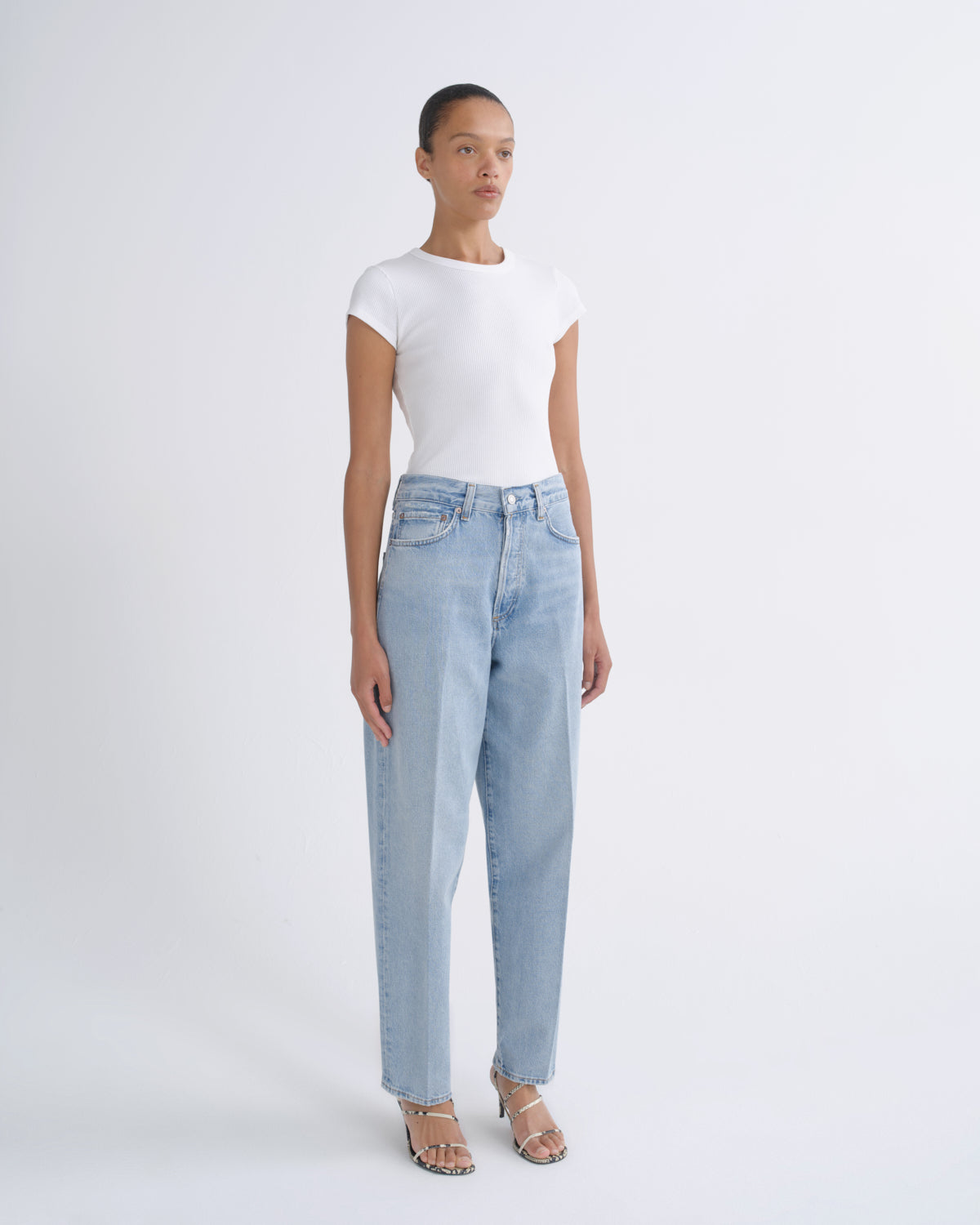 A model stands against a light gray studio background wearing a fitted white short-sleeve top, light-wash blue straight-leg jeans, and beige strappy sandals.