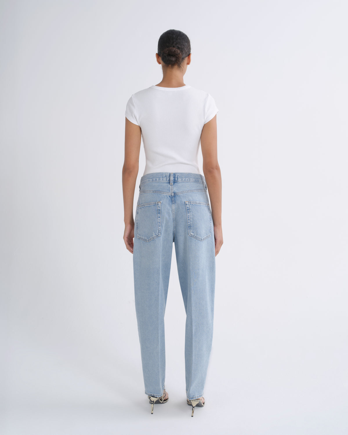 A model viewed from the back wearing a white short-sleeve t-shirt and light-wash blue jeans, standing against a plain light gray studio background.