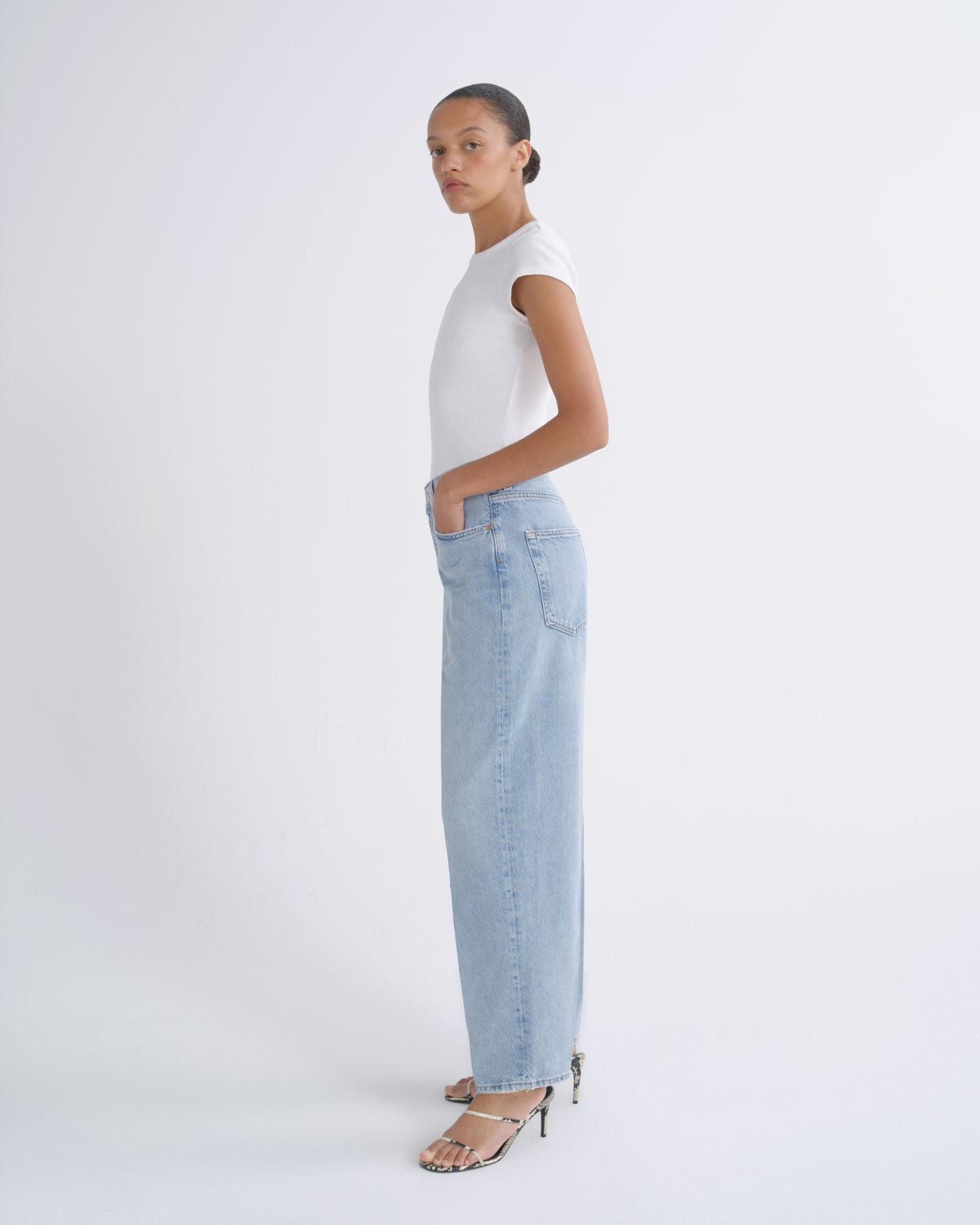 A model is shown in profile wearing a white sleeveless top, light-wash wide-leg denim jeans, and black strappy high-heeled sandals against a plain white studio background.