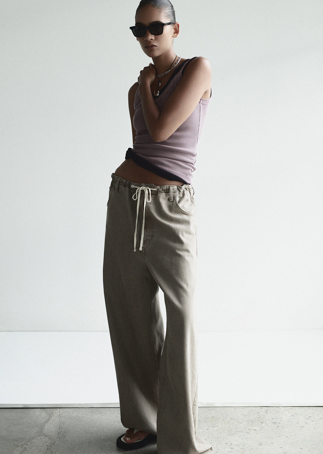 Person wearing a sleeveless top and beige pants with sunglasses on a plain background