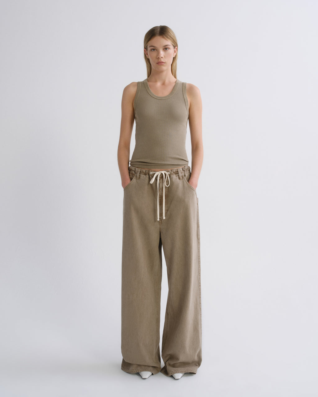 A woman stands against a light gray studio background wearing a sleeveless olive-beige tank top and matching wide-leg drawstring pants.
