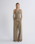 A woman stands against a light gray studio background wearing a sleeveless olive-beige tank top and matching wide-leg drawstring pants.