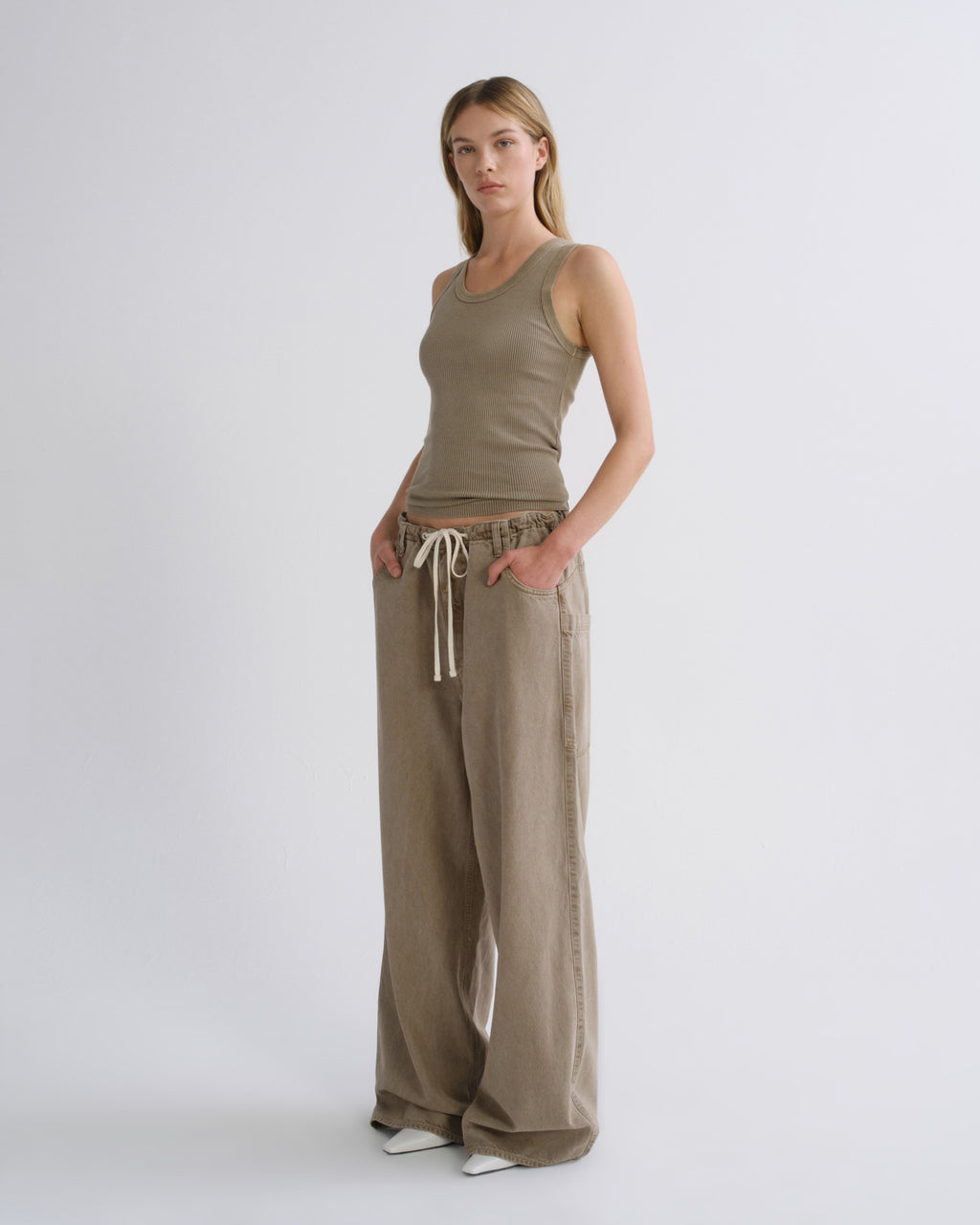 A woman modeling a matching two-piece lounge set in a light taupe color, featuring a ribbed tank top and wide-leg pants with a drawstring waist, set against a plain light gray studio background.
