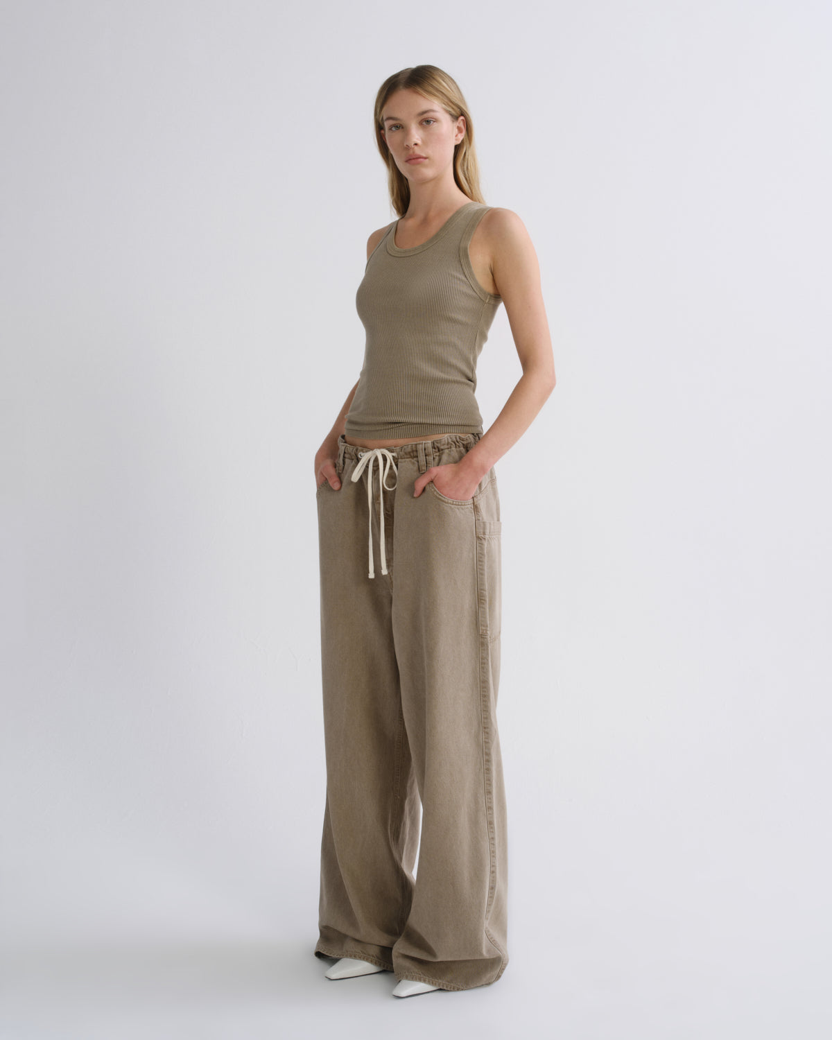 A woman modeling a matching two-piece lounge set in a light taupe color, featuring a ribbed tank top and wide-leg pants with a drawstring waist, set against a plain light gray studio background.