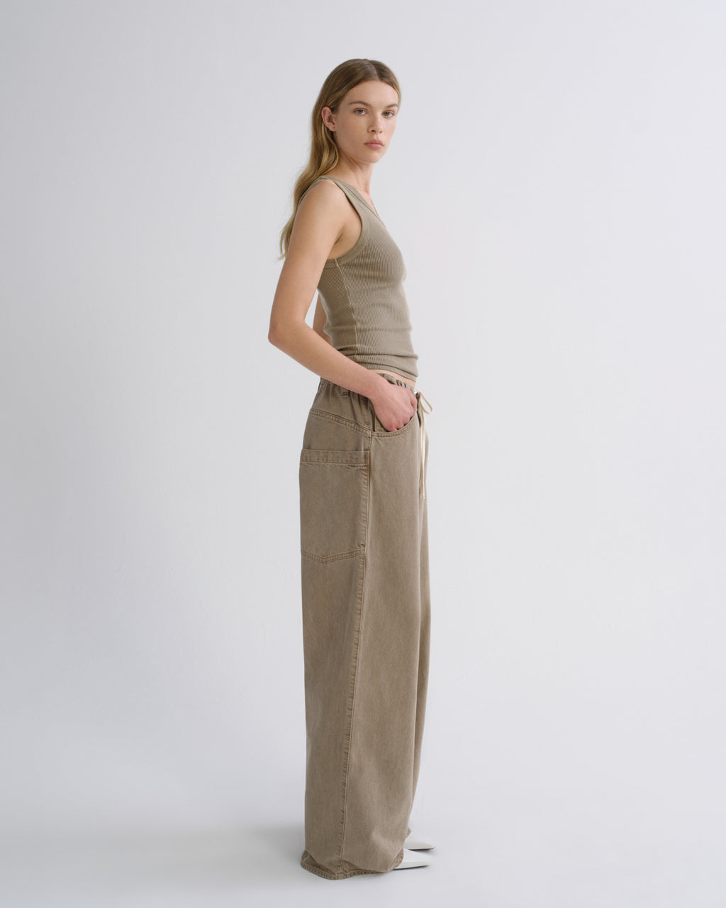 A woman stands in profile wearing a sleeveless taupe knit top and matching wide-leg trousers against a clean light gray studio background.