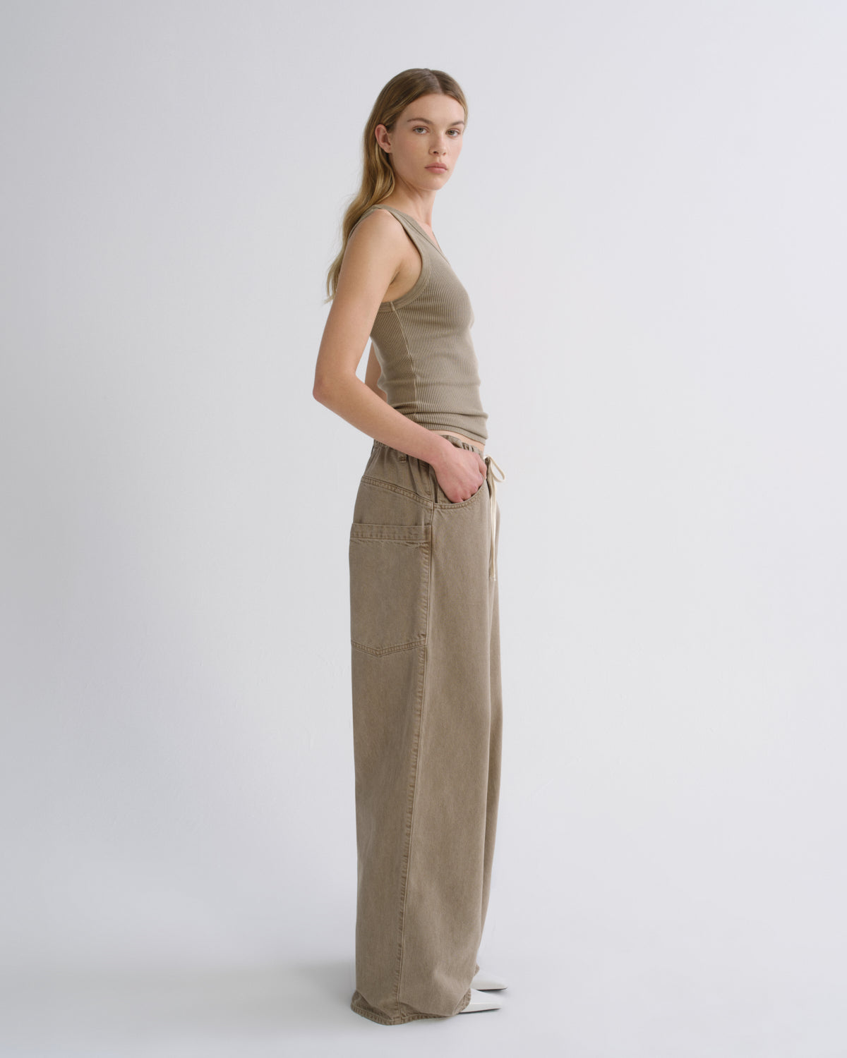 A woman stands in profile wearing a sleeveless taupe knit top and matching wide-leg trousers against a clean light gray studio background.