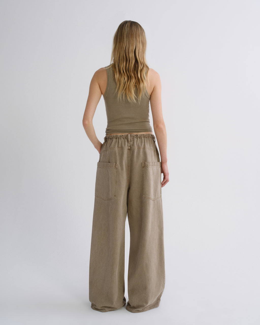A woman viewed from the back wearing a sleeveless taupe top and matching wide-leg pants with large patch pockets, standing against a light gray studio background.