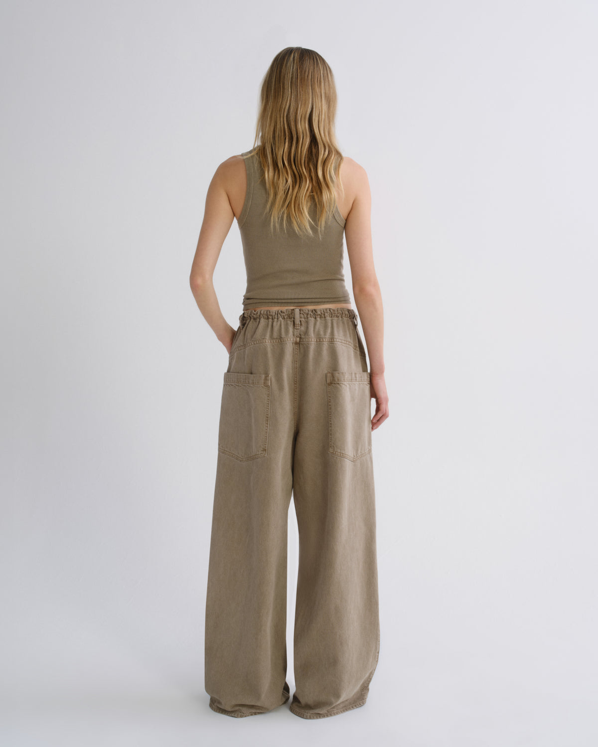 A woman viewed from the back wearing a sleeveless taupe top and matching wide-leg pants with large patch pockets, standing against a light gray studio background.