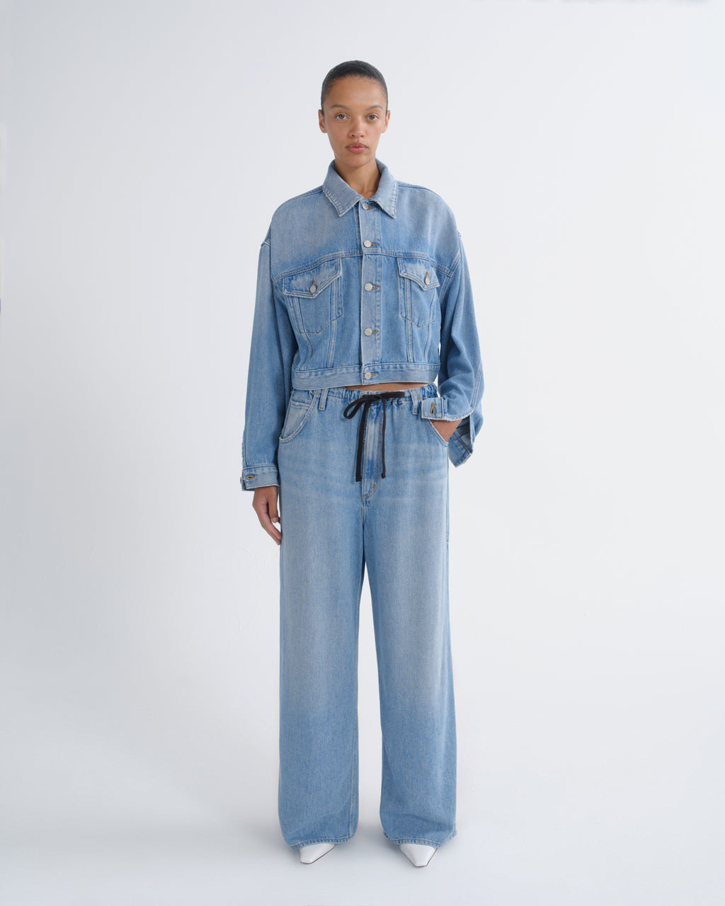 A model stands against a light gray studio background wearing a light blue denim set consisting of a cropped button-front jacket with long sleeves and matching wide-leg pants with a dark drawstring waist.