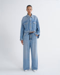 A model stands against a light gray studio background wearing a light blue denim set consisting of a cropped button-front jacket with long sleeves and matching wide-leg pants with a dark drawstring waist.