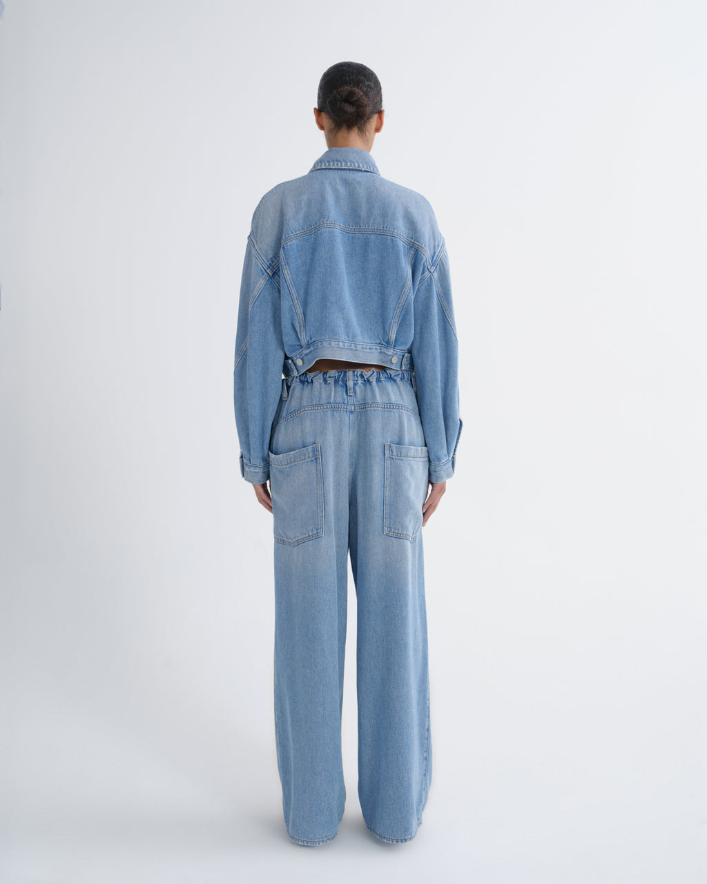 A model viewed from the back wearing a light blue denim jumpsuit with wide-leg pants, a cropped long-sleeve top, and a matching belt.