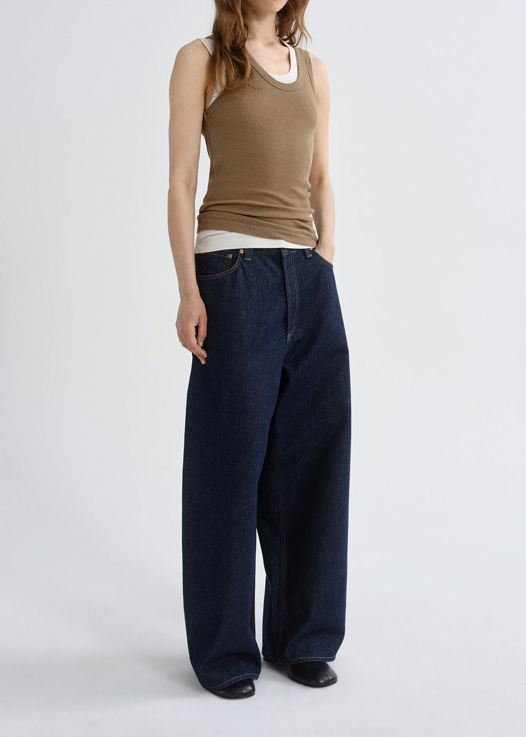 Person wearing a brown ribbed tank top with dark wash wide leg jeans viewed from the side on a grey background