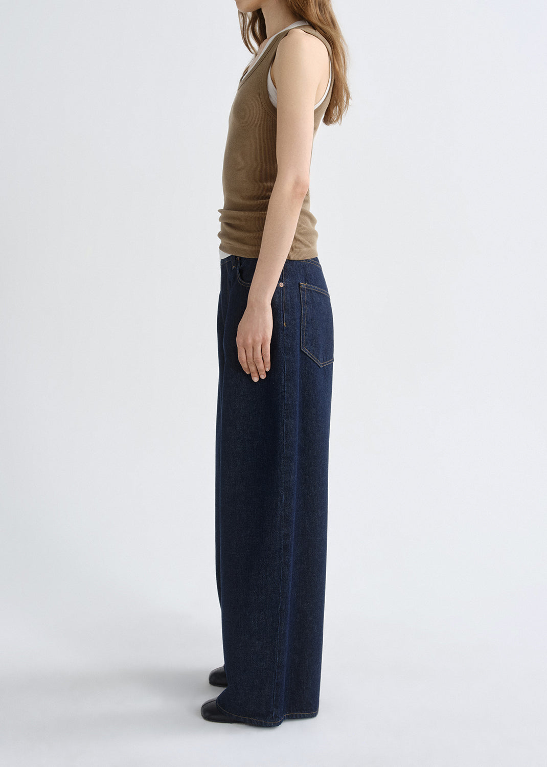 Person wearing a brown ribbed tank top with dark wash wide leg jeans viewed from the side on a grey background