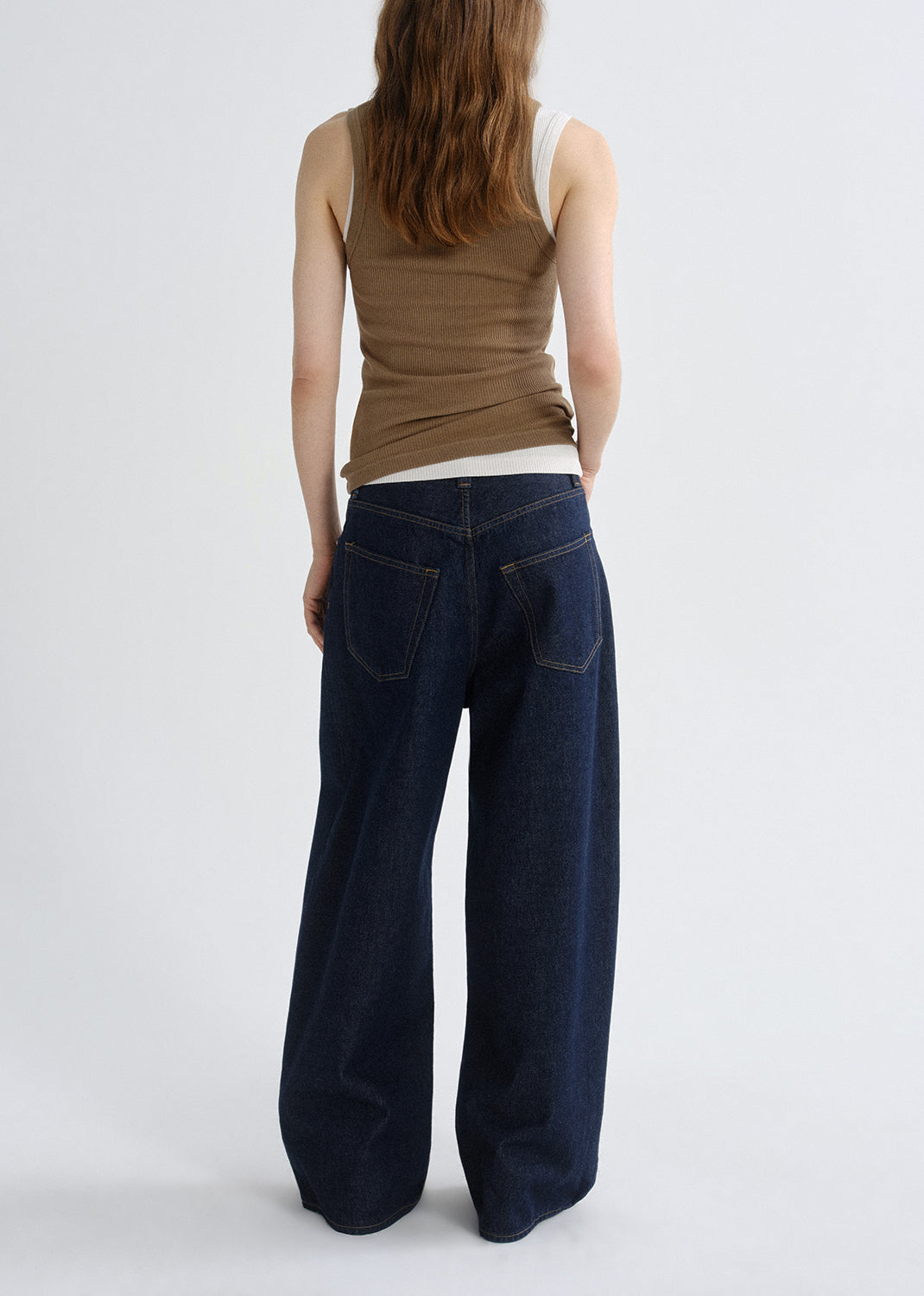 Person wearing a brown ribbed tank top with dark wash wide leg jeans viewed from the back on a grey background