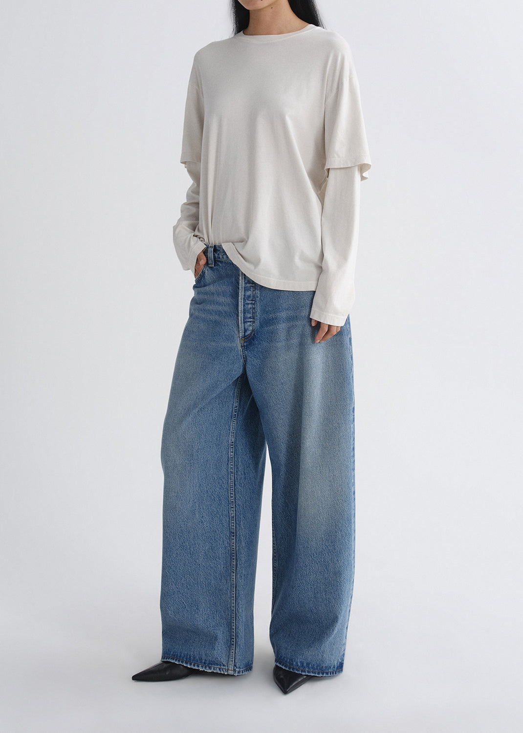 Person wearing a cream long sleeve tee with light wash wide leg jeans viewed from the side on a grey background