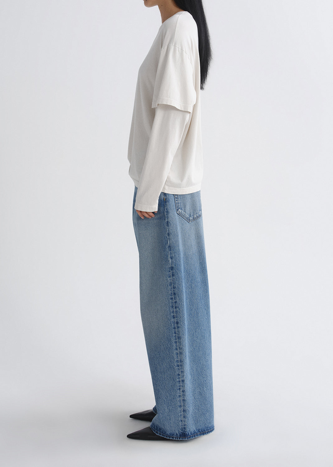 Person wearing a cream long sleeve tee with light wash wide leg jeans viewed from the side on a grey background