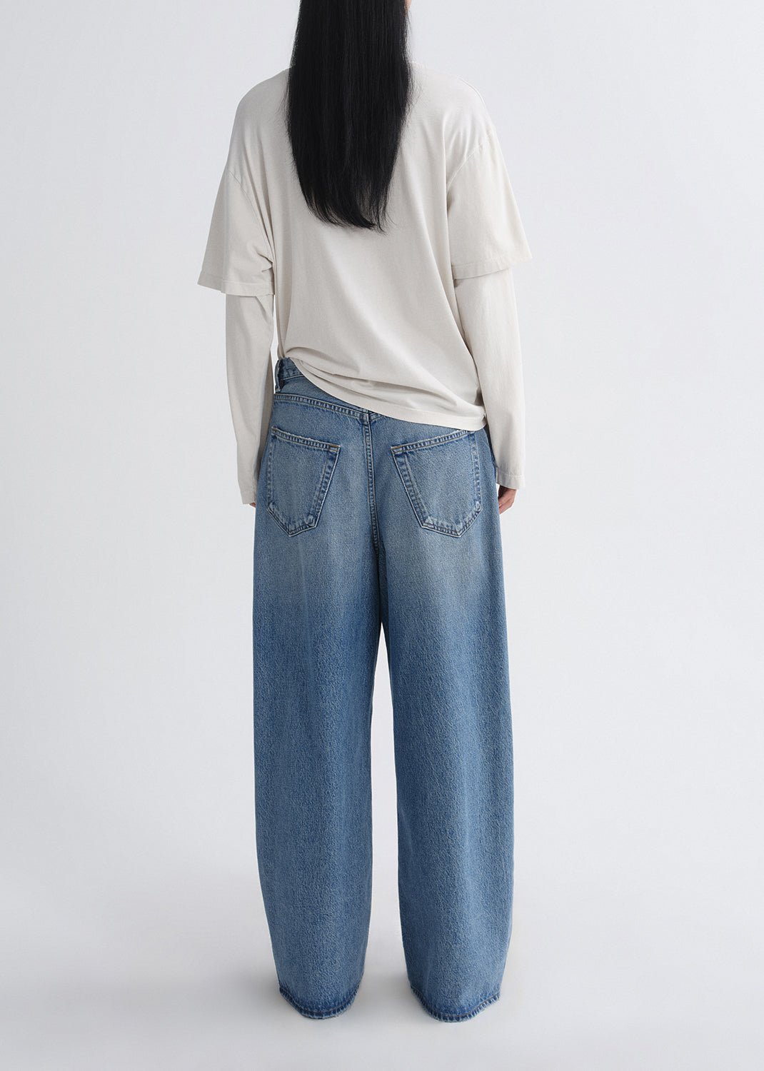 Person wearing a cream long sleeve tee with light wash wide leg jeans viewed from the back on a grey background