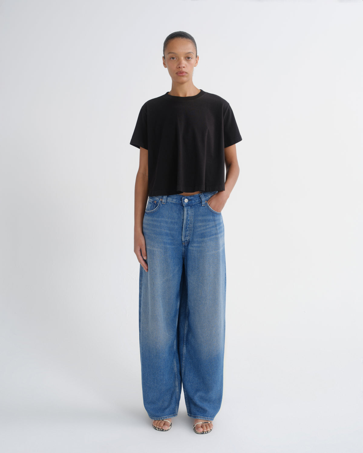 A model stands against a light gray studio background wearing a black short-sleeve cropped t-shirt and wide-leg blue denim jeans.