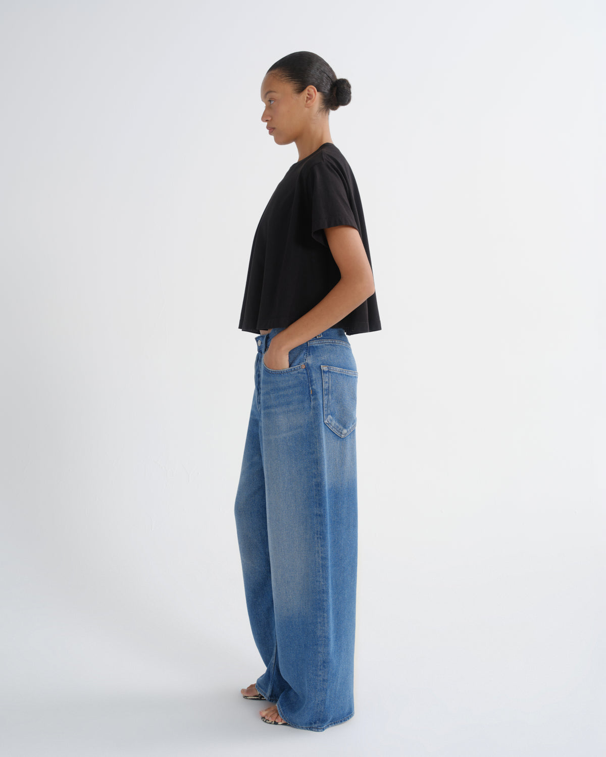 A model standing in profile wearing a black short-sleeve cropped t-shirt and wide-leg blue denim jeans against a plain white studio background.