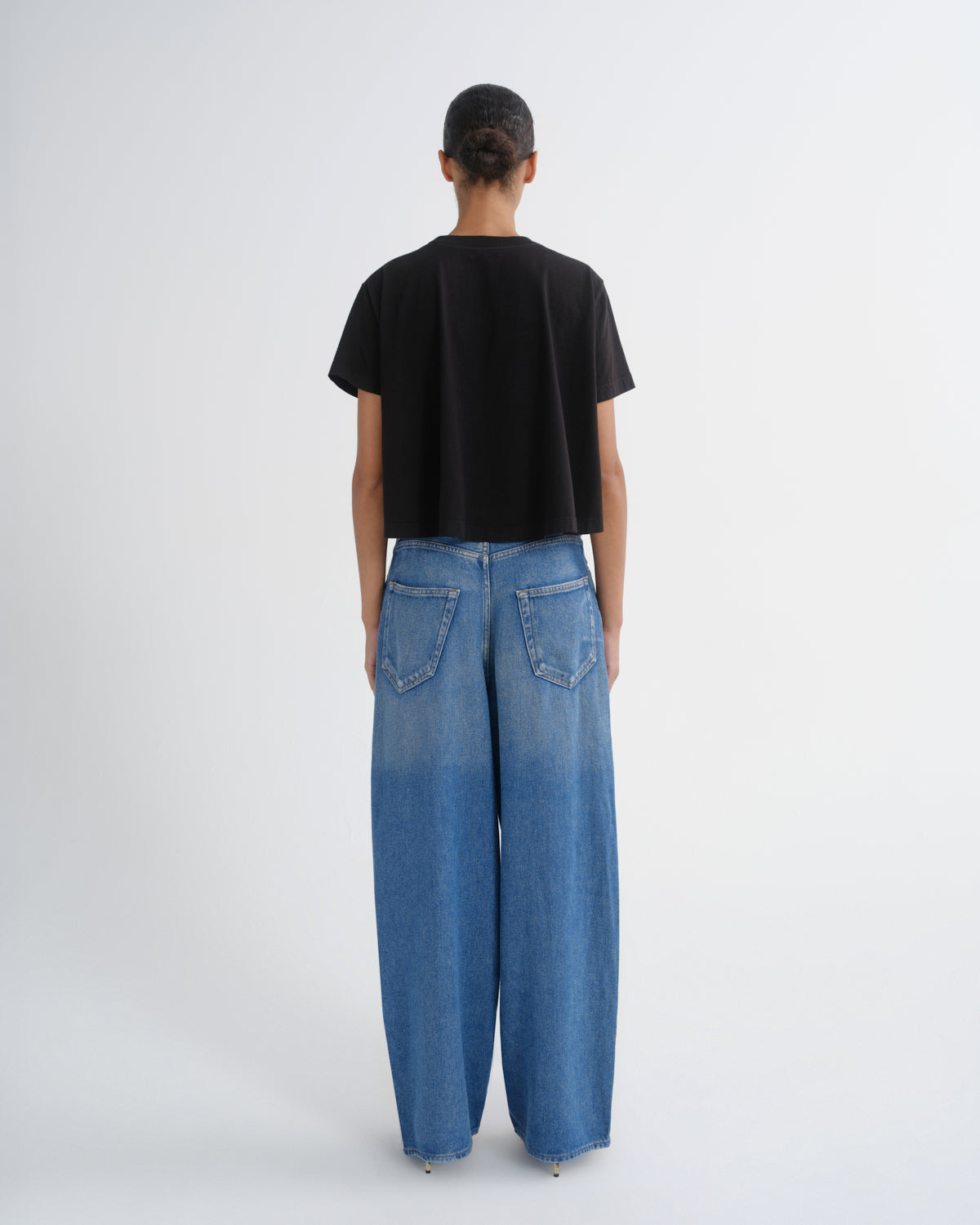 A model viewed from the back wearing a black short-sleeve crop top and wide-leg blue denim jeans against a plain light gray studio background.