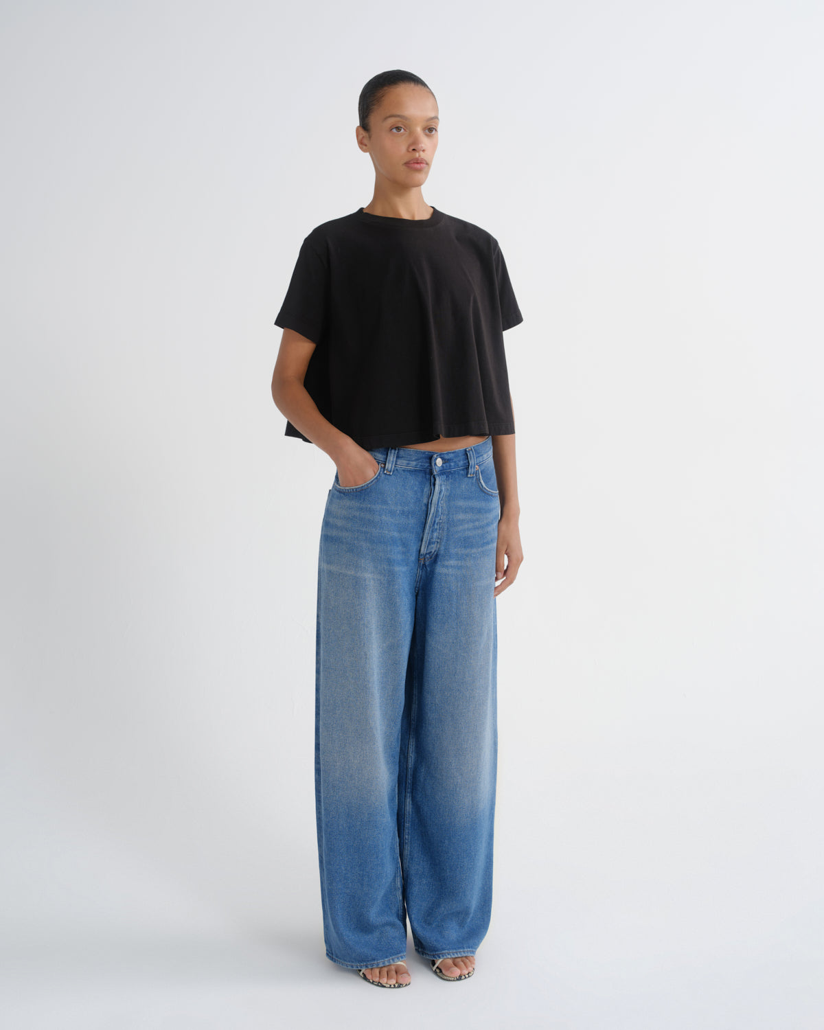 A model stands against a light gray studio background wearing a black cropped t-shirt and wide-leg blue jeans.