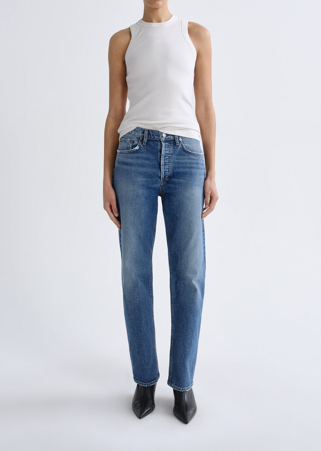 A model wearing a white sleeveless top and blue straight-leg jeans with black pointed-toe heels, standing against a grey background.
