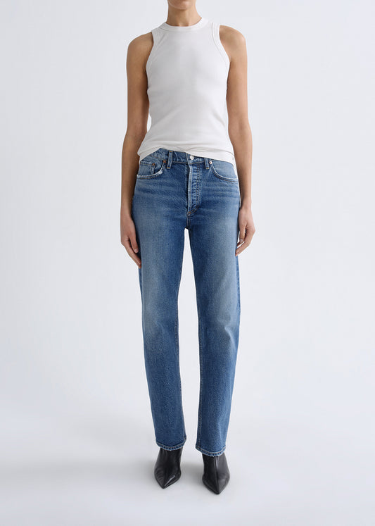 A model wearing a white sleeveless top and blue straight-leg jeans with black pointed-toe heels, standing against a grey background.