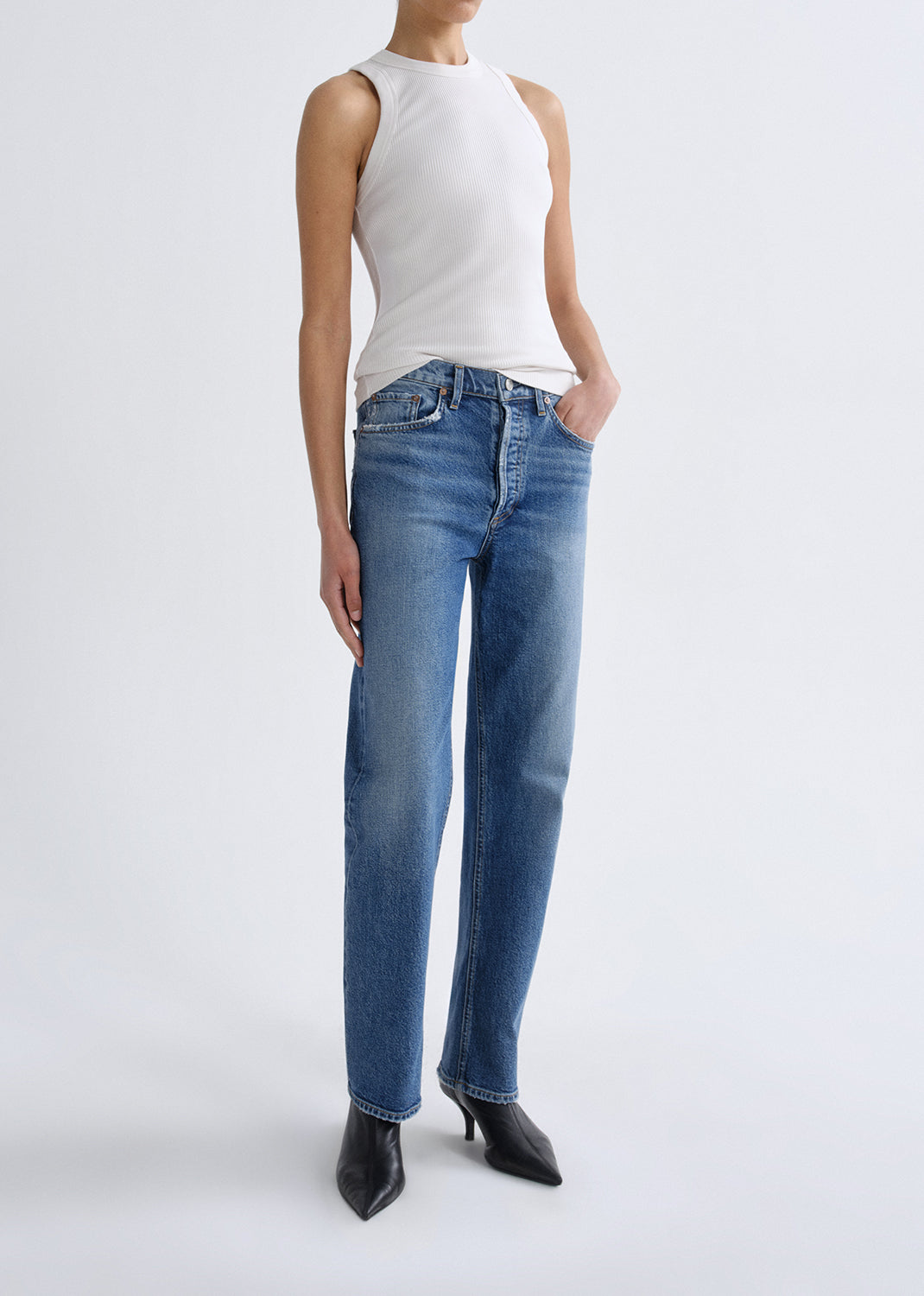 A model wearing a pair of medium-wash blue straight-leg jeans, paired with a white sleeveless top and black pointed-toe ankle boots, standing against a grey background.