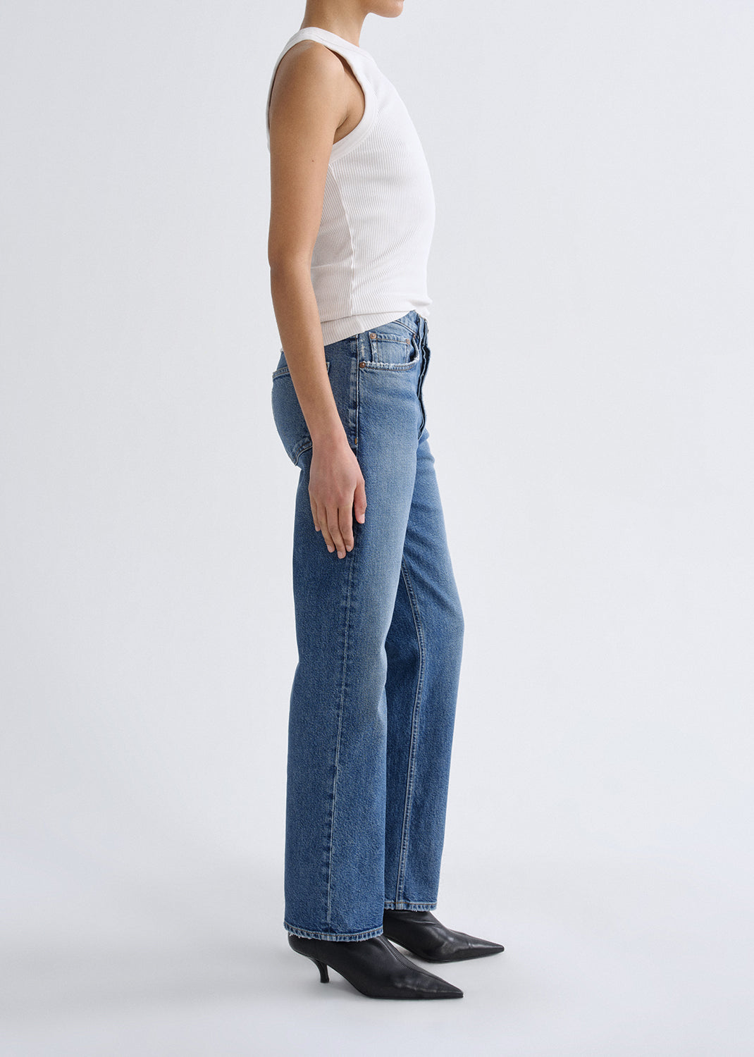 A side-profile studio shot of a model wearing blue straight-leg jeans, a white sleeveless top, and black pointed-toe heels against a grey background.